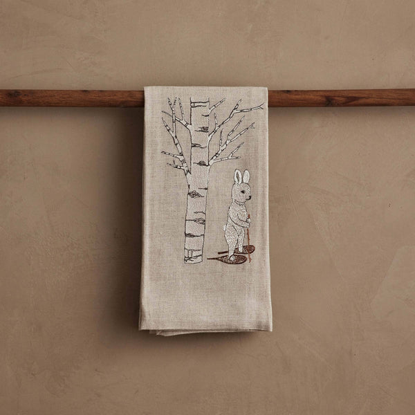 The Coral & Tusk Snowshoe Hare Tea Towel in beige linen hangs on a wooden rod, embroidered with a white rabbit beside a bare aspen tree for a serene winter scene.