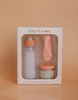 A boxed set called "My First Tiny Tummies gift Set - Milk and Cereal" includes a toy baby bottle, spoon, and a small jar of cereal, ideal for playtime. The scene is bathed in a soft, peachy hue, casting an enchanting glow over these tiny treasures.