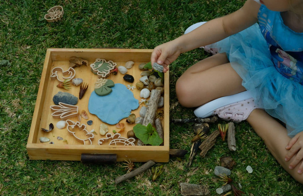 A child in a blue dress sits on the grass, playing with a wooden tray filled with clay shapes, small rocks, leaves, and sticks. Nearby, the Pond Life Cutter Set – an eco playdough tool made from biodegradable PLA – and an eco bubble wand add a sensory, earth-friendly touch to their nature-inspired play experience.