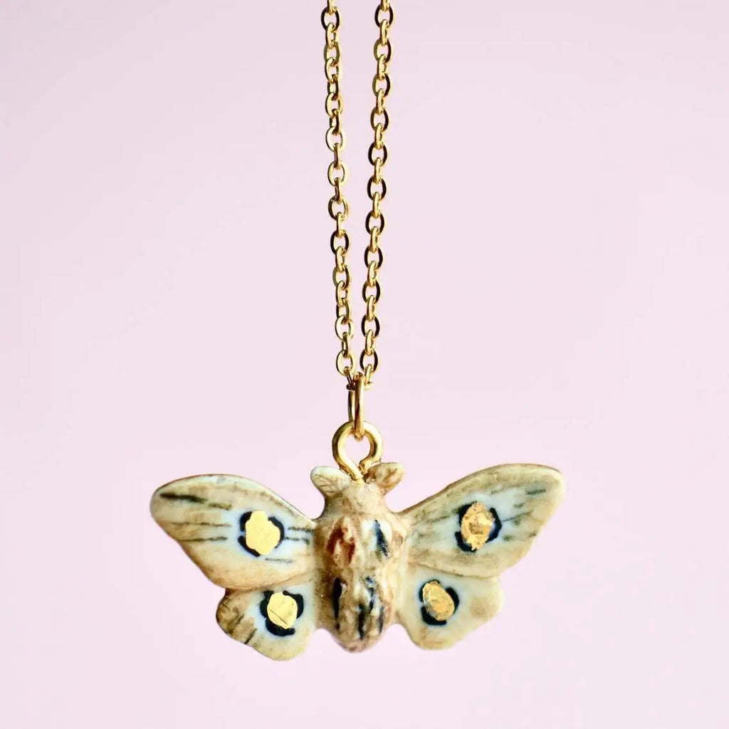 A Solar Moth Necklace with a delicate 24k gold plated chain, featuring yellow and black spots on its wings, set against a soft pink background.
