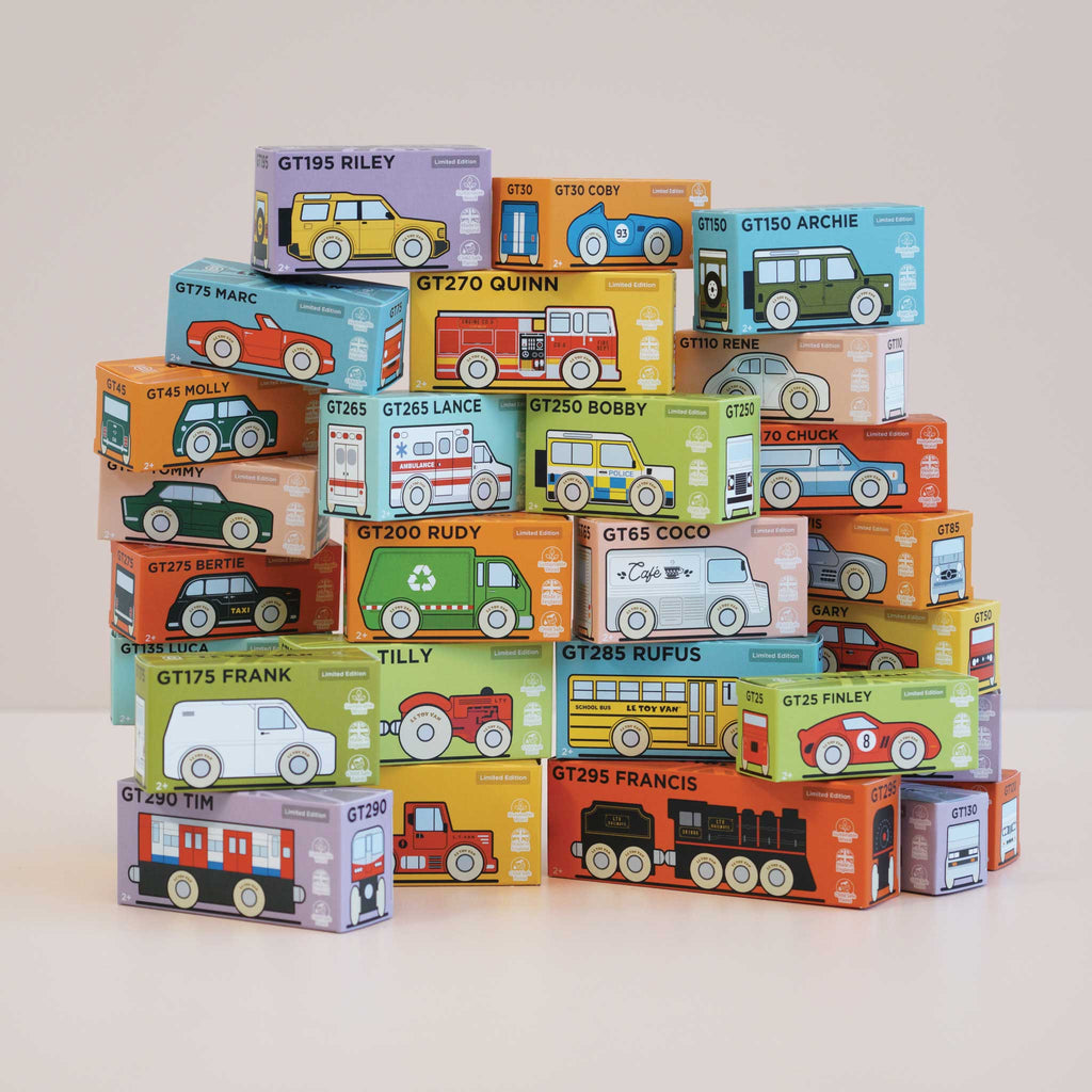 A pyramid of colorful toy vehicle boxes, each illustrated with cars, buses, and trains—including the No. 13 GT75 Marc, a classic toy wooden convertible in vibrant racing red made in England. The boxes are stacked neatly, displaying various names and designs against a plain white background.