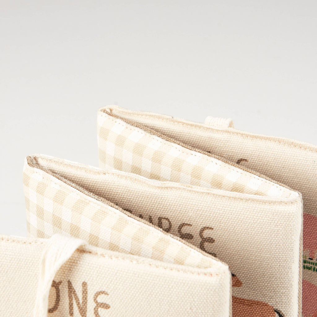 Close-up of three fabric-covered books, including the Little Farm Soft Book with beige and cream checkered and illustrated covers, fanned out on a white background—ideal as a baby gift.
