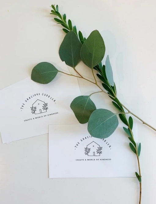 A sprig of eucalyptus and a stem with small leaves lie diagonally across a white surface, beside The Gracious Gobbler Bundle: Children's Book, Plush and Cards featuring the logo and motto "Gracious Gobbler - create