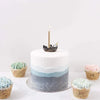 A white cake with blue ombre layers on a plate, decorated with a single lit candle and two Sea Otter Cake Toppers on top, surrounded by multicolored frosted cupcakes.
