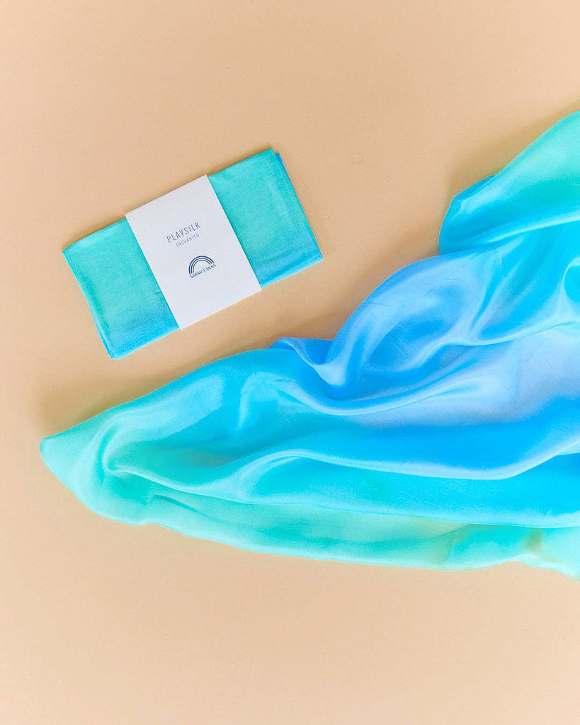 A folded Sarah's Silks Enchanted Playsilk - Sea, wrapped in a white band, rests next to a loosely draped matching scarf on a light beige background, inviting open-ended play.