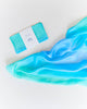 A folded Sarah's Silks Enchanted Playsilk - Sea with a white label sits beside its unfurled, smooth gradient aqua and turquoise silk, shown on a white surface—perfect for open-ended play.