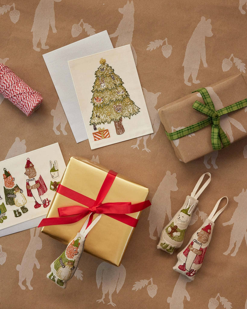 A selection of holiday-themed items on brown paper adorned with animal silhouettes: greeting cards, gifts wrapped with red and green ribbons, and two North Pole Bear Ornaments. The cards display a Christmas tree along with festive characters.