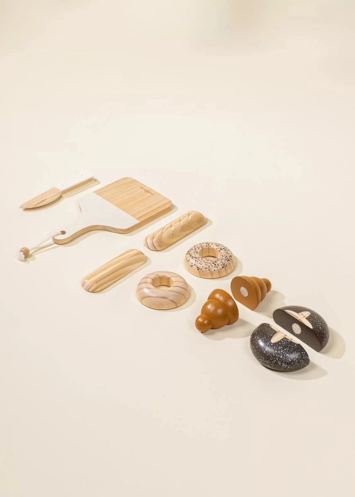 The Wooden Bakery Set, perfect for children's games, includes a toy cutting board, knife, croissant, baguette, doughnut, and various types of bread. The toys have a light wood finish and are displayed on a neutral background.