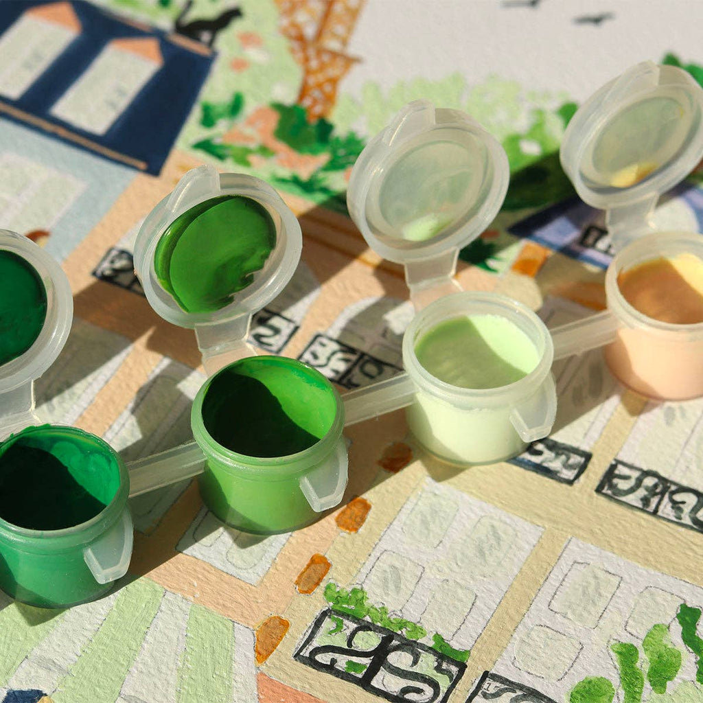 Small open paint pots in green and cream shades rest on a partially painted Paint By Number Kit - Paris illustration of sunlit buildings and trees, inviting aspiring artists to express their creativity.