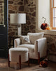 A cozy nook with a white armchair, ottoman, and side table is accented by plush pumpkins and the Coral & Tusk Bat Wing Surprise Pillow by the window on a stone wall, adding warm autumn charm.