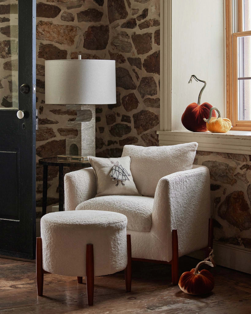 A cozy nook with a white armchair, ottoman, and side table is accented by plush pumpkins and the Coral & Tusk Bat Wing Surprise Pillow by the window on a stone wall, adding warm autumn charm.