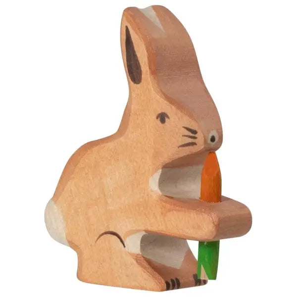 A small wooden figurine of a bunny holding an orange and green carrot. Crafted from handcrafted maple and beech wood, the bunny is carved in a simple, minimalist style with painted details for the eyes, nose, and fur markings. This Holztiger Rabbit With Carrot is light brown with darker accents on the ears and feet. Made in Europe.

