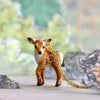 A miniature porcelain figurine of a Fawn Necklace, with a 24k gold plated chain attached to its neck, standing amid a soft-focus natural backdrop with rocks.