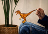 A person in jeans holds a Schleich Velociraptor toy with a movable jaw, sitting next to a woven plant pot with tall green leaves.