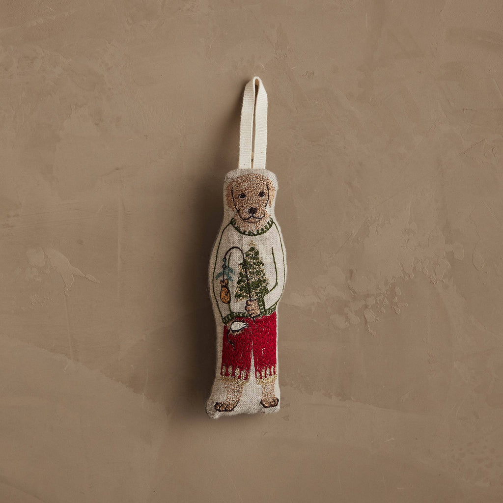 The Coral & Tusk Dog with Gifts Ornament features a bear in a festive dog sweater and red pants, holding a tree and lantern, hanging by a fabric loop against a textured beige wall.
