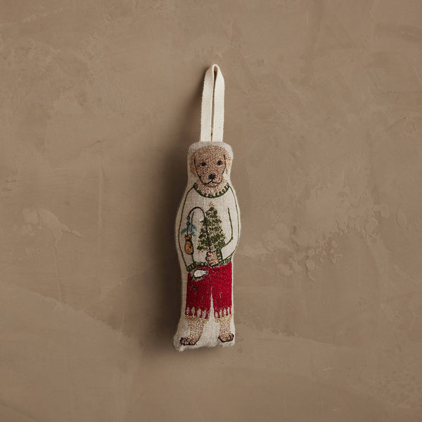 The Coral & Tusk Dog with Gifts Ornament features a bear in a festive dog sweater and red pants, holding a tree and lantern, hanging by a fabric loop against a textured beige wall.