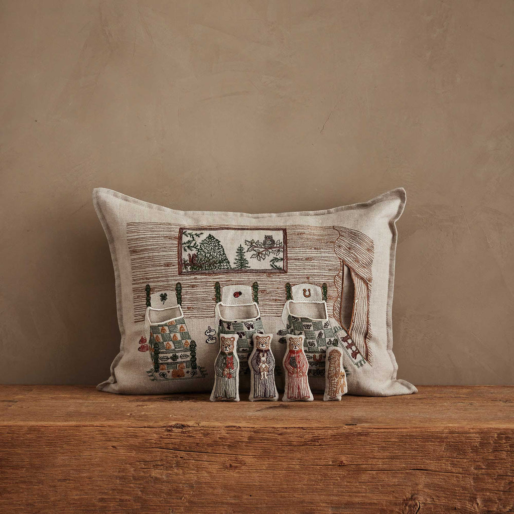 The Coral & Tusk Three Little Bears Pocket Pillow, featuring an embroidered cottage scene, sits against a tan wall with five charming, decorative cat figurines arranged on a rustic wooden surface. Ships in one week.
