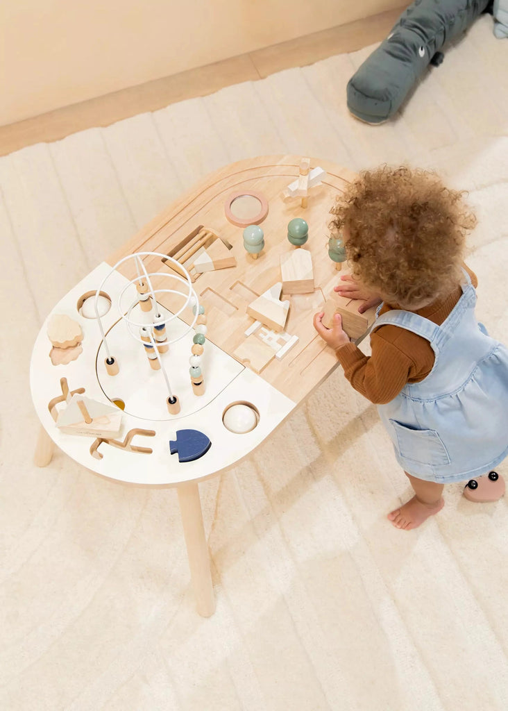 A toddler with curly hair stands next to a Wooden Activity Table shaped like a kidney, filled with various birch plywood toys, including blocks and a bead maze, in a warmly lit room.