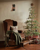 A cozy living room features a brown armchair with a green blanket and the Christmas Donkey Pillow beside a festively decorated Christmas tree with wrapped gifts underneath, all aglow in warm, inviting lighting.