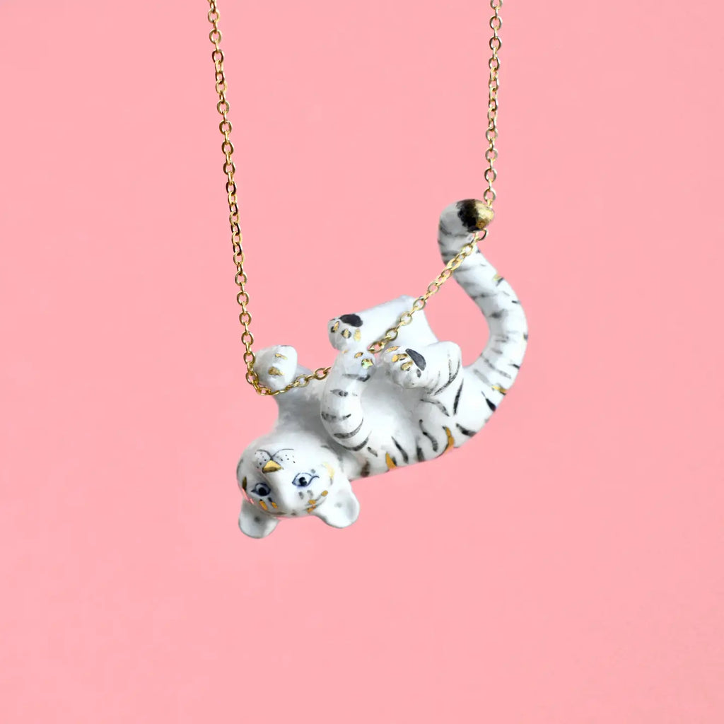 A delicate Gold Tiger Necklace featuring a small, playful white tiger pendant with black stripes, depicted hanging upside down, grasping the 24k gold plated steel chain with its paws. The hand-painted porcelain charm stands out beautifully against the solid pink background, highlighting the charming design.