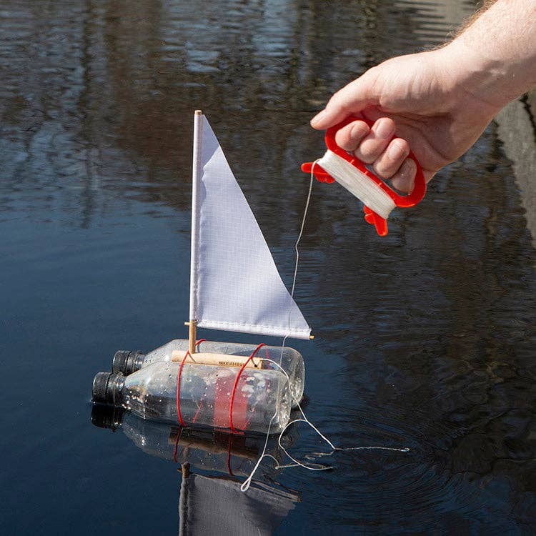 The Huckleberry Make Your Own Sailboat, featuring a white sail, floats on water as a hand holds the attached string.