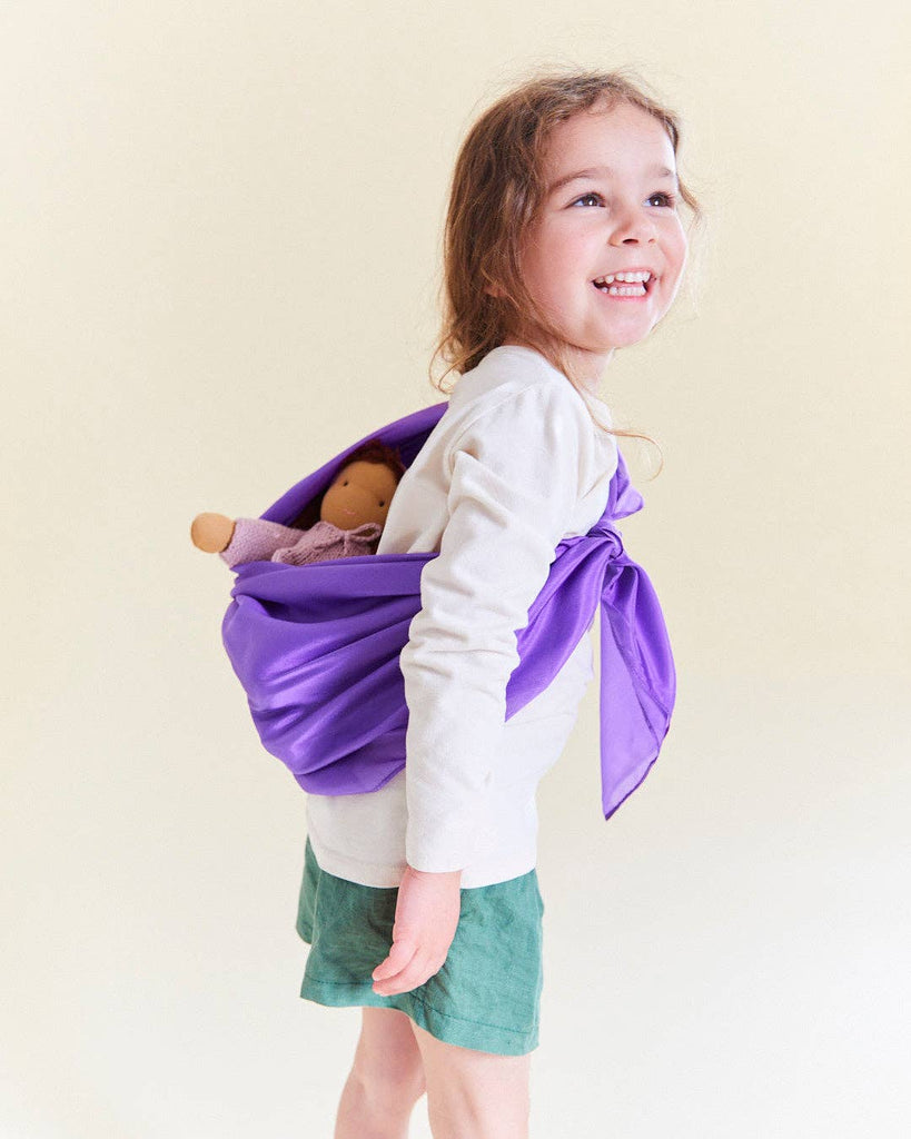 A smiling young girl in a white shirt and green shorts carries a doll in a Sarah's Silks Solid Color Playsilk - Purple sling over her shoulder, inspiring open-ended play against a light background.