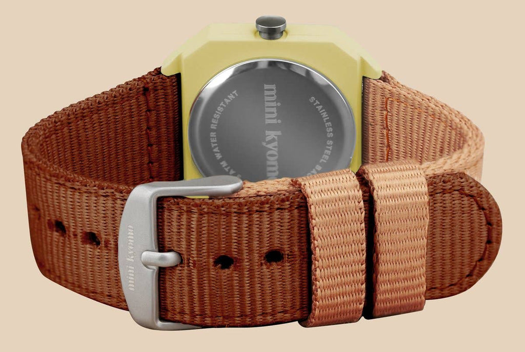 A close-up of the Mini Kyomo Watch - Cherry Bomb shows a brown fabric strap, stainless steel back, “mini kyomo” on the clasp, and a light yellow case that enhances its vintage charm.