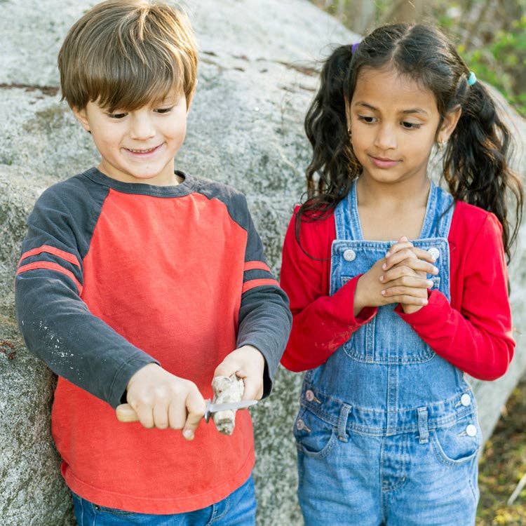 A young boy in a red shirt carves a rock with the Huckleberry Carving Knife (Not a Toy) as a girl in denim overalls and a red shirt watches, both standing outside near a large stone.
