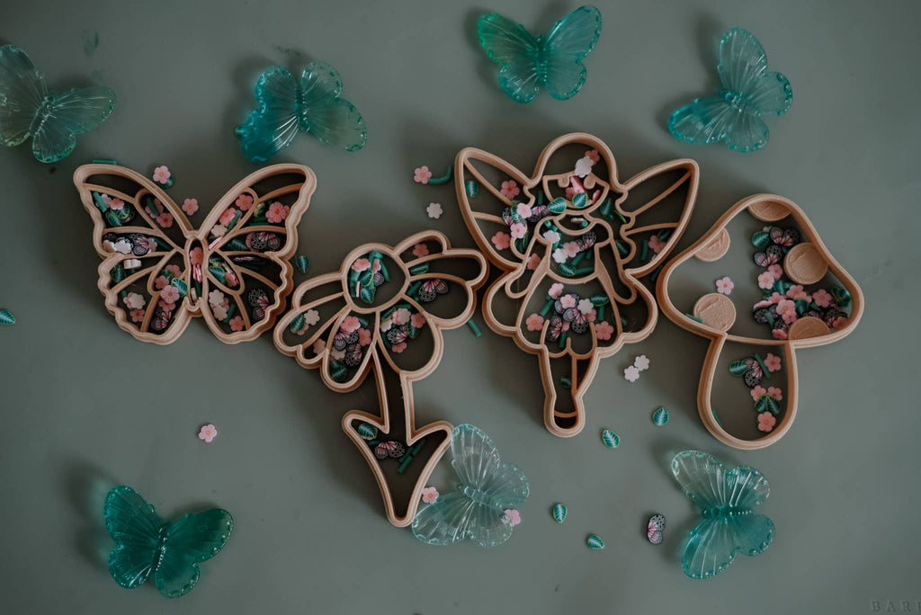Butterfly, flower, fairy, and mushroom-shaped trays filled with small floral pieces are arranged on a surface, surrounded by translucent blue butterfly decorations — a perfect setup for sensory play using the Fairy Cutter Set - Eco Playdough Tool or your favorite fairytale-themed tools.