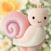 A hand holds the Cuddle + Kind Baby Snail (Peony), a hand-knit plush toy with a pink and lavender spiral shell, cream body, black eyes, smiling mouth, and brown antennae, set against blurred yellow and white flowers.