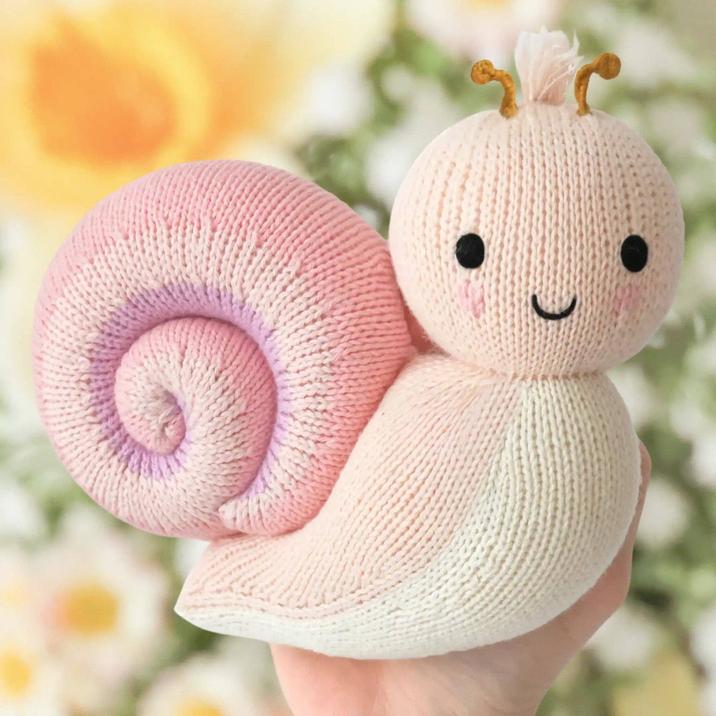A hand holds the Cuddle + Kind Baby Snail (Peony), a hand-knit plush toy with a pink and lavender spiral shell, cream body, black eyes, smiling mouth, and brown antennae, set against blurred yellow and white flowers.