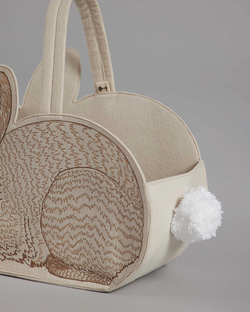 A Coral & Tusk Bunny Basket handbag with embroidered details and a fluffy white pom-pom tail, photographed against a gray background.