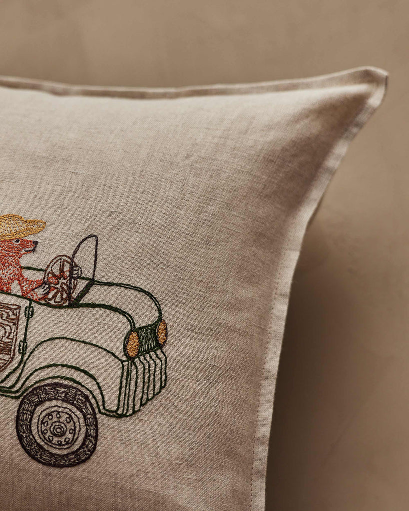 The Coral & Tusk Christmas Tree Car Pocket Pillow is a beige linen pillow with embroidered art of a bear in a hat driving a vintage green car, partially visible on the left side.