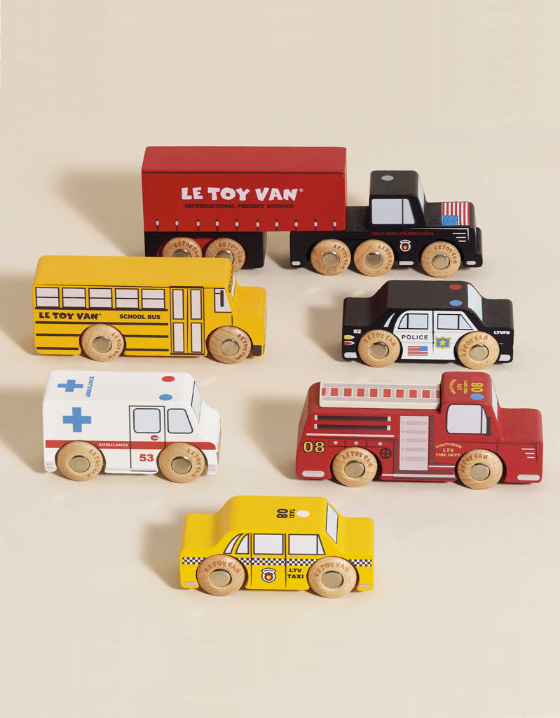 The American Wooden Car Set features six colorful vehicles—a red truck, yellow school bus, police car, ambulance, fire truck, and yellow taxi—perfect for imaginative play and displayed on a light beige surface.