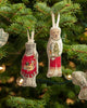 The Coral & Tusk Dog with Gifts Ornament hangs from a green Christmas tree adorned with lights and festive decorations in the background.