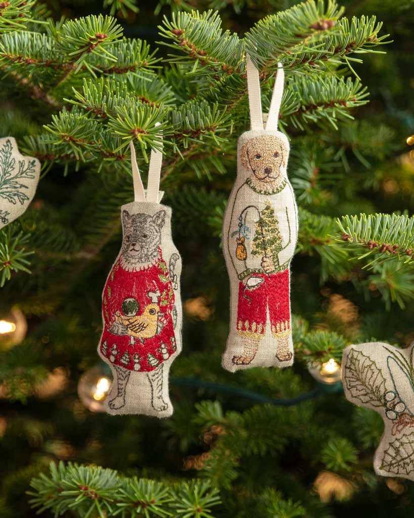 The Coral & Tusk Dog with Gifts Ornament hangs from a green Christmas tree adorned with lights and festive decorations in the background.