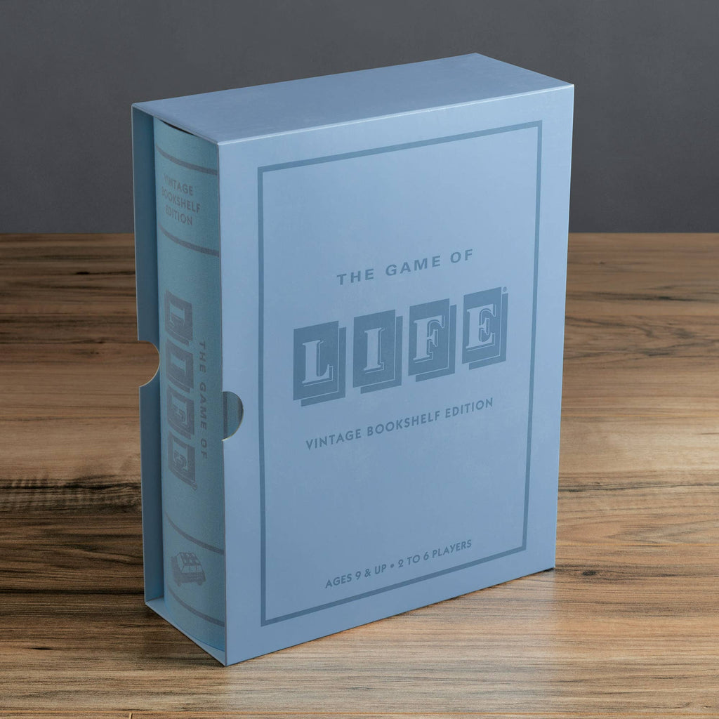 A light blue book-shaped box labeled "Board Game: The Game of Life Vintage Bookshelf Edition" rests on a wooden surface. This classic game is ideal for ages 9+, perfect for 2 to 6 players.