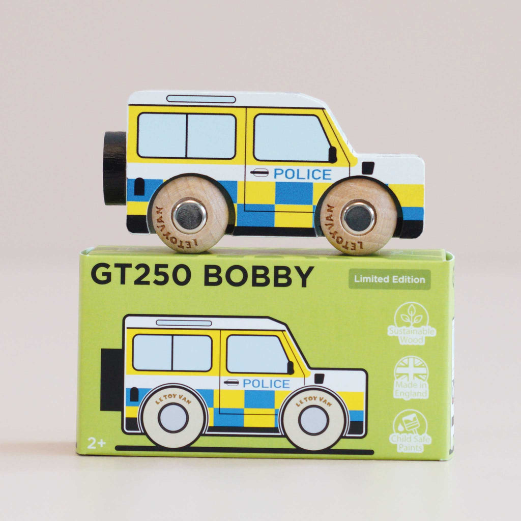 A wooden police car with blue and yellow checkered details, inspired by classic British patrol vehicles, is displayed atop a green box labeled “No. 40 GT250 Bobby | Made in England.” The box features an illustration of the police car alongside eco-friendly icons—making it perfect for imaginative play.