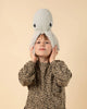 A young child stands against a beige background, dressed in a leopard-print top. They are wearing a grey, knitted hat shaped like a BigStuffed Mini Grandma Octopus, with the octopus's large eyes visible near the top of the head. The child holds the sides of the hat with both hands.