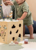 A baby in green clothes plays with the Wooden Activity Cube, enjoying bead mazes and shape sorters to develop fine motor skills on a soft mat. An adult sits nearby, partially visible in the background.
