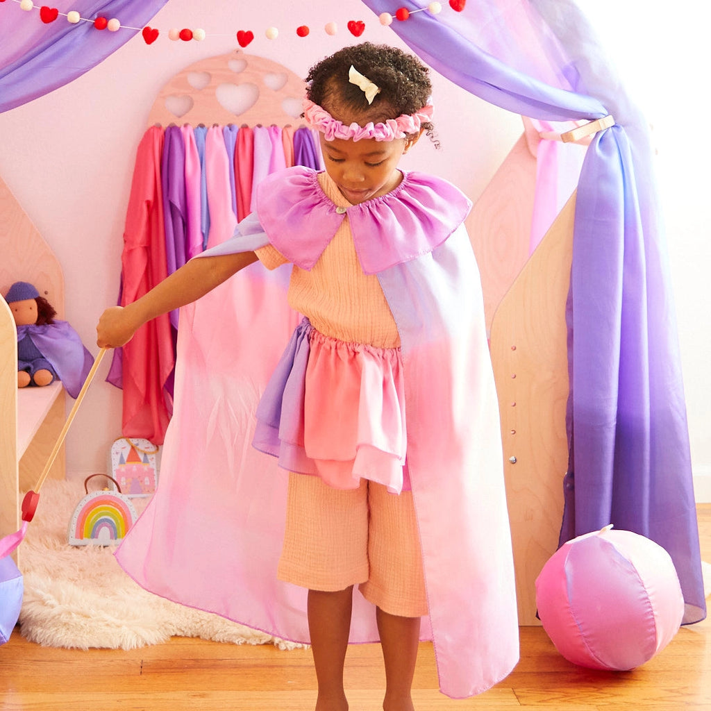 A child dressed in a colorful costume enjoys some dress-up play in a vibrant play area. The ensemble includes a purple and pink cape, Sarah's Silk Blossom Garland Headband, and peach-colored outfit. Behind the child, garlands hang from a fabric canopy with vibrant drapes and toys.