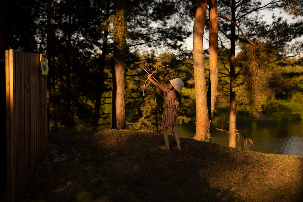 A young boy in a straw hat and striped outfit practicing archery by a lake at sunset, aiming with a Wooden Bow & Arrows Set with Goal at a target beside a wooded area.