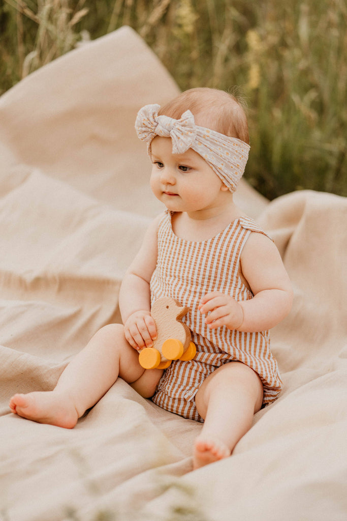 A baby in a striped romper and headband sits on a beige blanket outdoors, holding the Sabo Concept Rolling Duck - Yellow. Grass and greenery can be seen in the background.