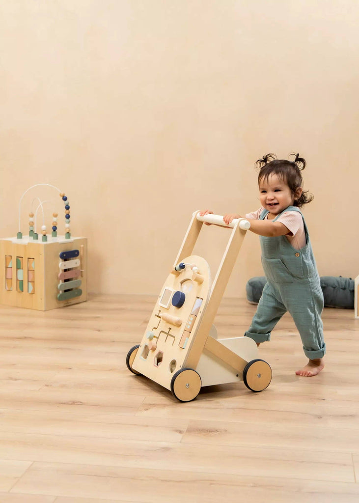 A toddler wearing blue overalls pushes the Wooden Activity Walker across a light wood floor. In the background, there is a wooden activity cube and a gray cushion.