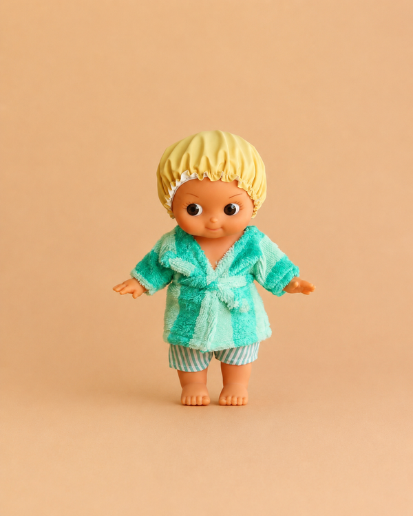 The Minikane Mini Pancha Doll - Basile with Green Striped Bath Set features a big-eyed doll dressed in a yellow shower cap, green robe, and striped shorts, standing barefoot on a beige background—perfect for imaginative play.