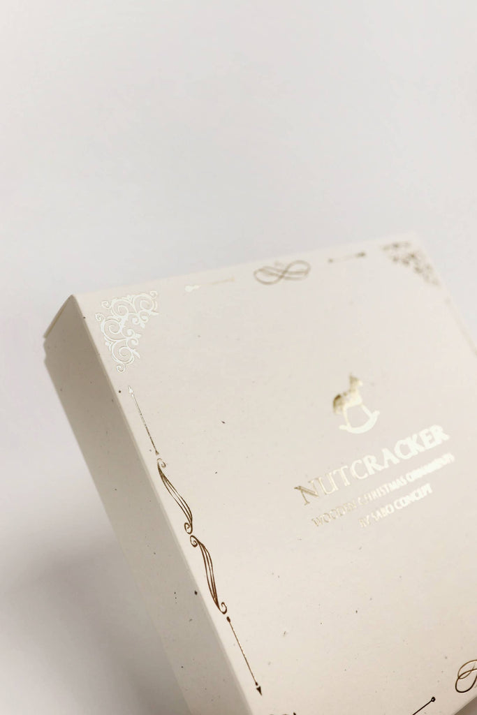 A close-up of the elegant Sabo Concept Christmas Ornaments box, cream-colored with gold designs and "Nutcracker" text on the lid, showcasing hand-painted wooden nutcracker ornaments against a plain white background.