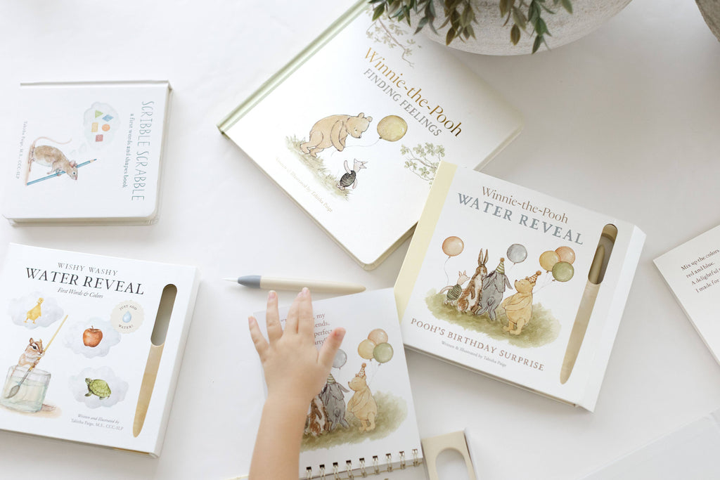 A child’s hand reaches for a Winnie-the-Pooh: Pooh’s Birthday Surprise (Water Reveal) activity book, surrounded by several other Winnie-the-Pooh storybooks and a stylus pen.