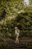 A young child with the Children's Backpack - Turtle, featuring adjustable shoulder straps, stands on a forest path. Sunlight filters through the trees, adding to the playful, adventurous mood as they look back over their shoulder.
