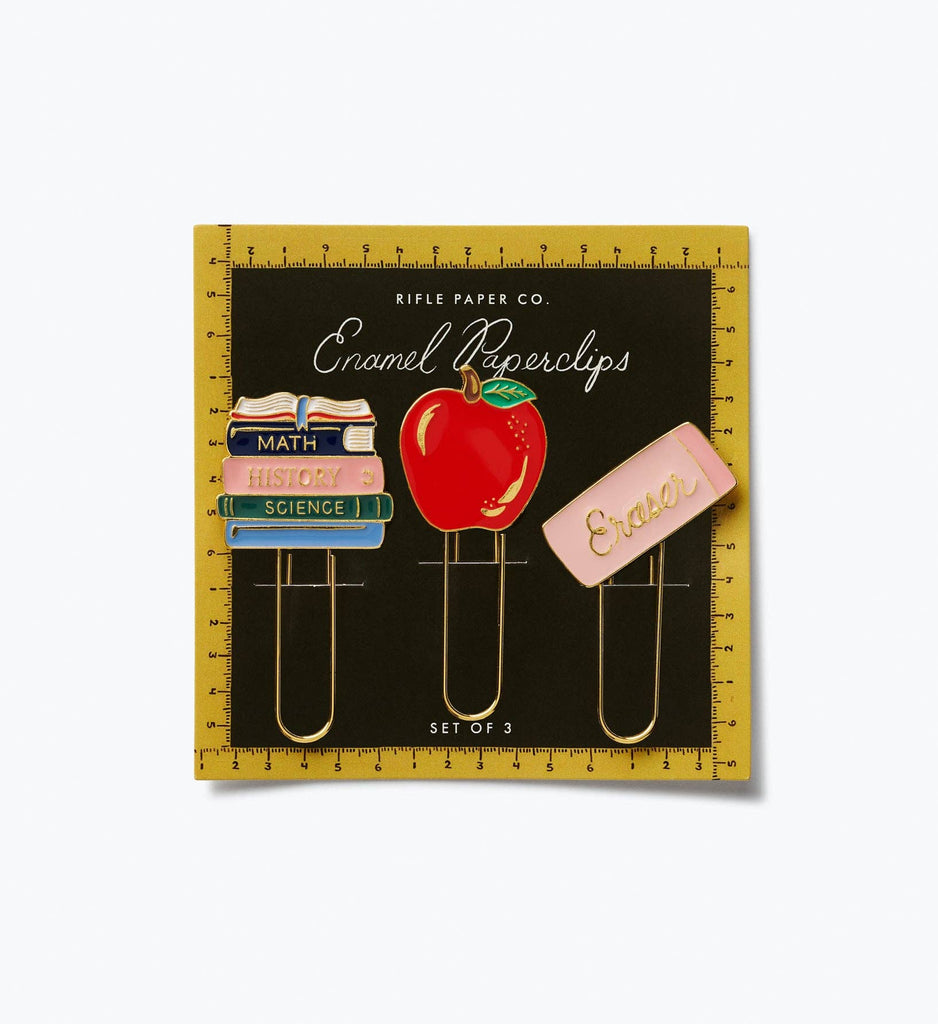 The Schoolhouse Enamel Paper Clip Set includes three oversized enamel paper clips: one featuring a stack of books labeled “Math,” “History,” and “Science,” one with a red apple, and one with a pink eraser, all presented on a black and yellow card.