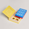 A yellow, book-shaped box for the Board Game: Guess Who? Vintage Bookshelf Edition lies open, showing a classic game board with blue and red frames and two illustrated Mystery Cards.